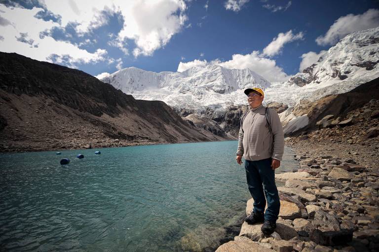 Homem de pé na margem de um lago de cor azul com montanhas cobertas de neve ao fundo