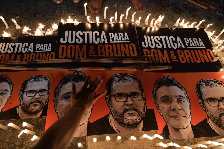 Cartazes com os rostos de Bruno e Dom e com o escrito 'Justiça para Dom e Bruno' estão dispostos no chão cercados por velas acesas; um mão posiciona um dos cartazes