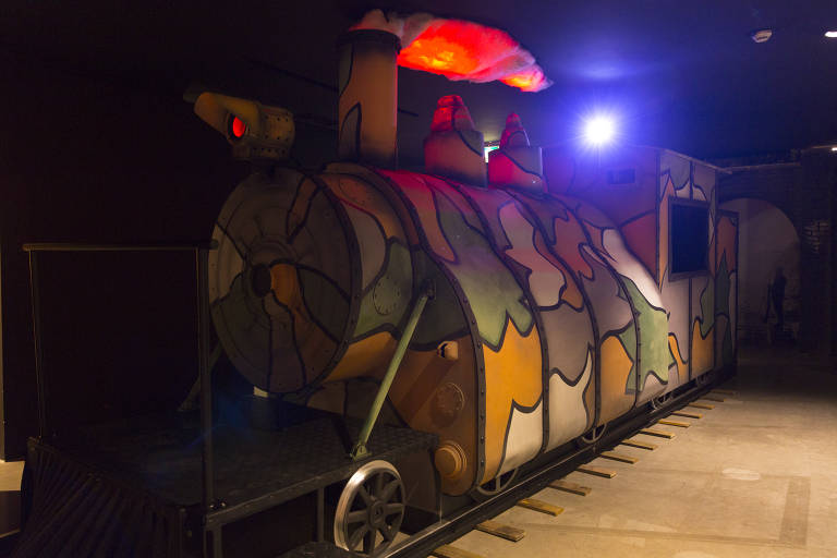 A imagem mostra a reprodução de um trem em tamanho real, pintado nas cores amarela, verde, branca e roxa. Uma luz sai da cabine do maquinista