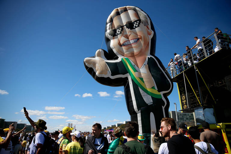 Boneco inflável gigante de Bolsonaro, fazendo sinal de arminha com a mão, é erguido por manifestantes ao lado de um carro de som