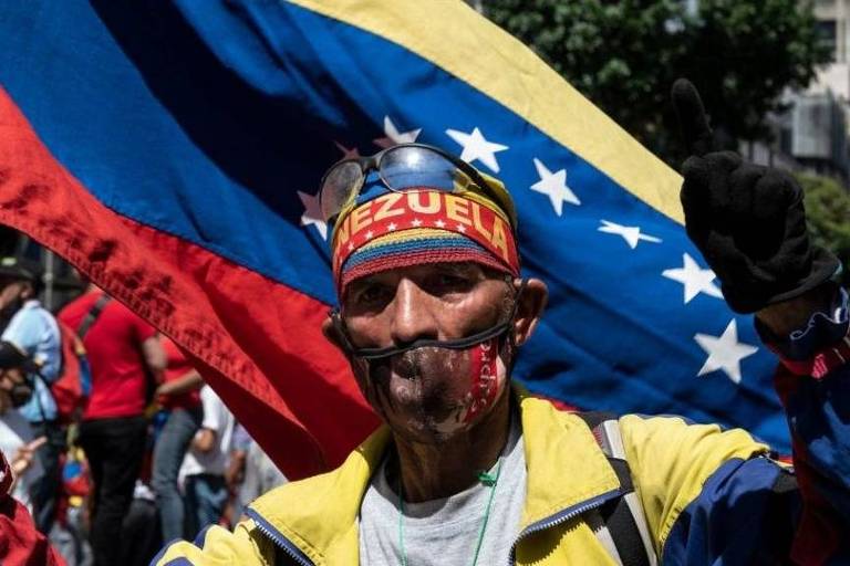 Imagem mostra um homem com uma bandeira da Venezuela ao fundo; ele usa também uma faixa com o nome do país na cabeça 