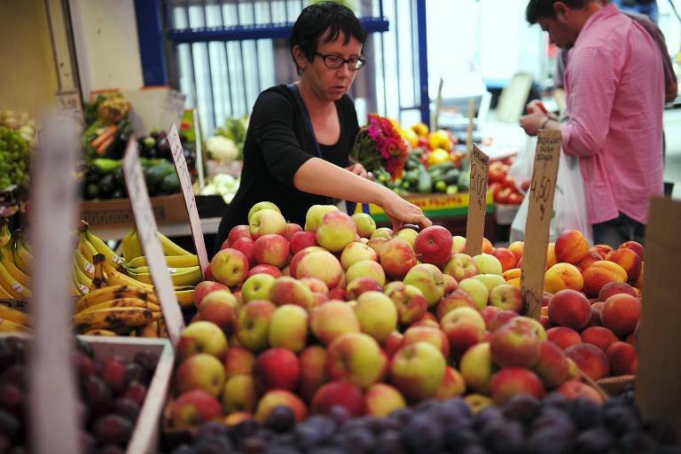 Imagem mostra mulher em hortifruti cercada de bancas com frutas. Ela está escolhendo algumas maçãs. Ao lado dela, há um momem de costas.