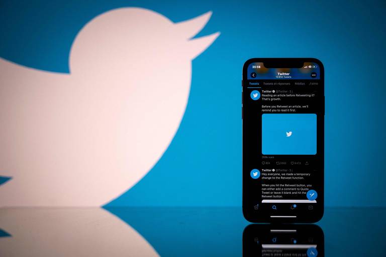Celular com Twitter aberto, com imagem do logo do Twitter (pássaro branco sobre fundo azul). Ao fundo, logo do Twitter.