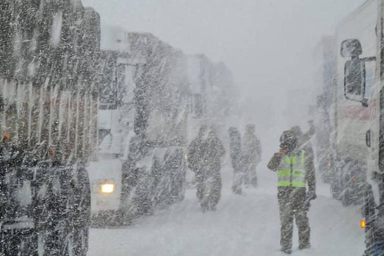 Vultos de caminhões são vistos em uma rodovia durante uma tempestade de neve; o chão está todo coberto por gelo