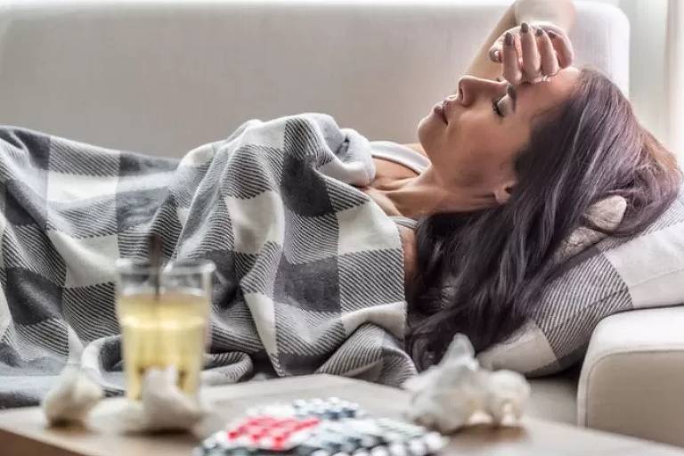 Imagem em primeiro plano mostra uma mulher deitada em um sofá coberta com uma manta. Ao lado, há uma mesa com remédios
