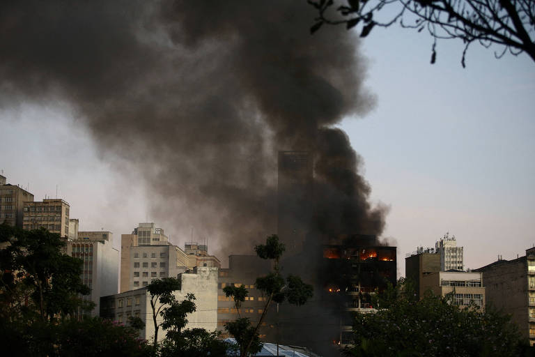 Imagem mostra prédio pegando fogo e fumaça preta subindo a um céu de final de tarde. Em volta do incêndio há cerca de uma dúzia de outros prédio. Em primeiro plano, é possível ver galhos e folhas de árvores.