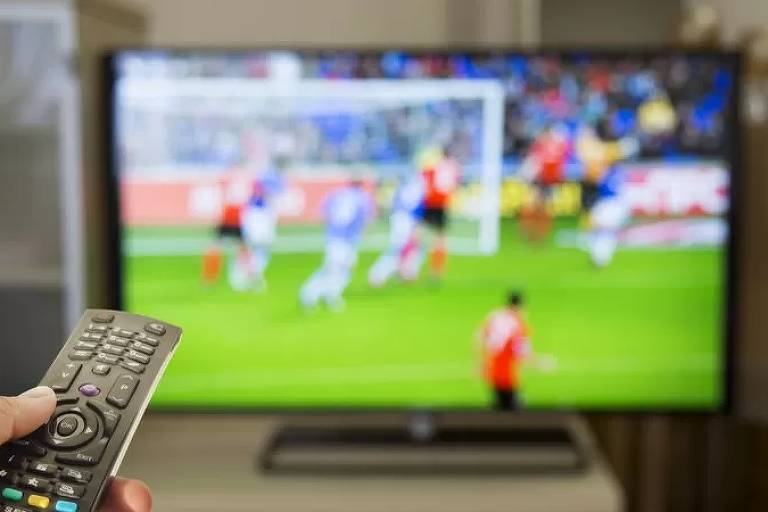 Imagem mostra a tela de uma televisão, onde passa uma partida de futebol. No canto esquerdo da imagem, se vê uma mão segurando um controle remoto