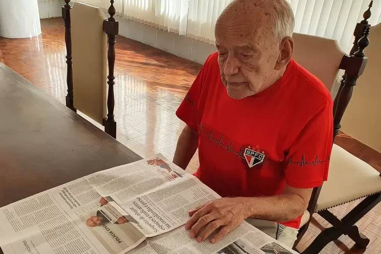 Imagem em primeiro plano mostra um homem idoso sentado de frente para uma mesa folheando um jornal