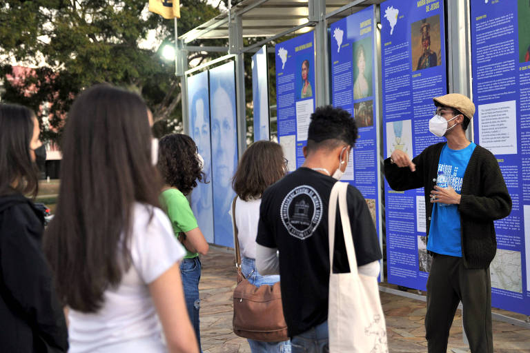 No canto da imagem, um homem com a camiseta azul da exposição fala e gesticula enquanto jovens observam, do outro lado