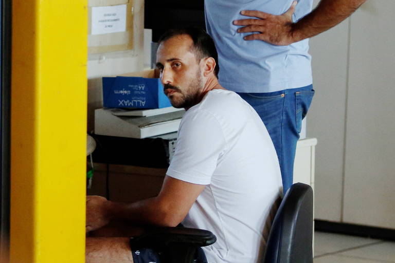 Giovanni Quintella está sentado e olha para trás, em direção à porta, de onde a foto é tirada; ele usa camiseta e bermuda