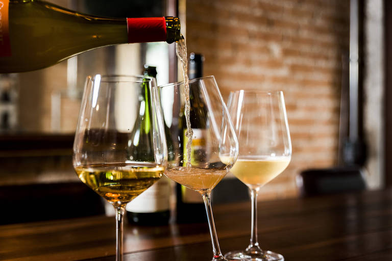Imagem mostra três taças de vinho branco uma ao lado da outra sobre um balcão de maneira. A primeira taça está 1/4 completa com vinho translucido, a segunda está sendo completada com um vinho cuja garrafa aparece na parte superior da imagem e a terceira está 1/4 completa com um vinho branco parcialmente opaco. 