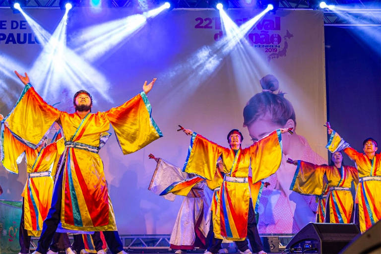 A foto mostra um grupo de artistas vestindo quimonos coloridos em um palco