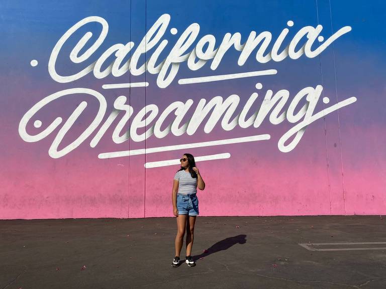 Fotografia colorida é retrato de Larissa em frente a um mural em que se lê "California Dreaming"