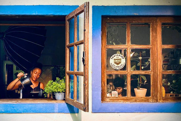 A pesquisadora Patty Durães coa café frente a uma janela aberta em uma fachada de uma casa azul