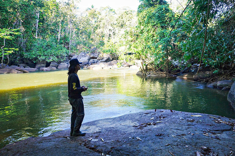 Na beira do leito de um rio, em meio à floresta, um jovem usa moletom e segura controle nas mãos; perto dele, está voando um drone