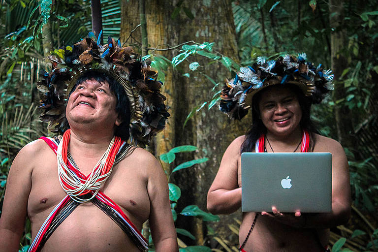 Um homem indígena olha em direção ao alto, enquanto, ao lado, uma mulher indígena segura um MacBook, notebook da marca Apple