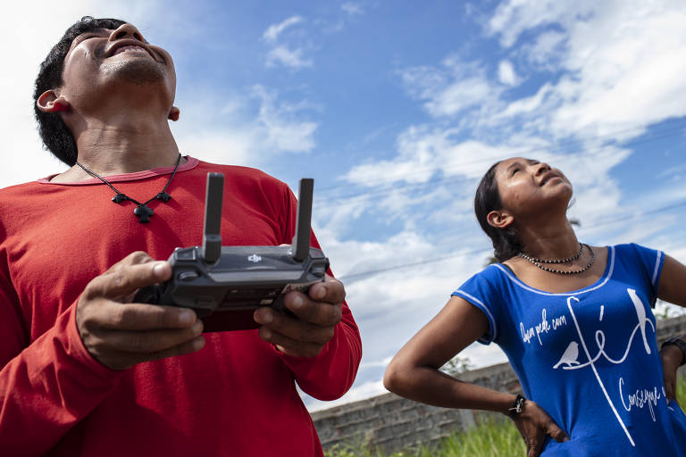 Dois jovens olham para o céu; um deles segura um controle com antenas, que comanda drone