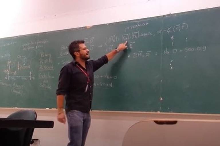 Imagem em primeiro plano mostra um homem em pé em uma sala de aula, na frente de uma lousa. Ele olha e aponta o dedo indicador para a lousa