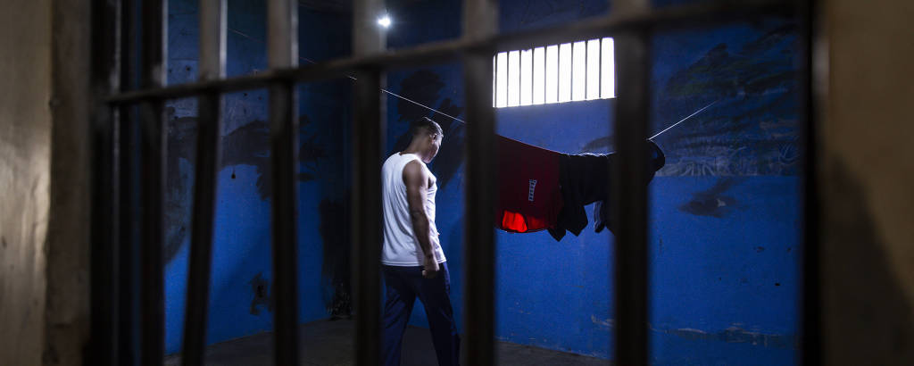 Homem vestido com camiseta branca e calça azul caminha dentro de uma cela escura com um varal de roupas, em frente a uma pequena janela por onde entra luz. Grades estão em primeiro plano