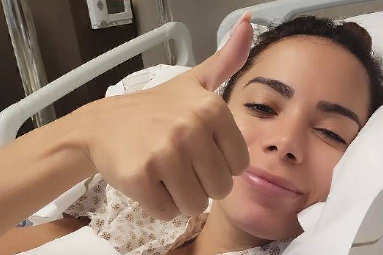 Em foto colorida, mulher faz sinal de positivo na cama de um hospital