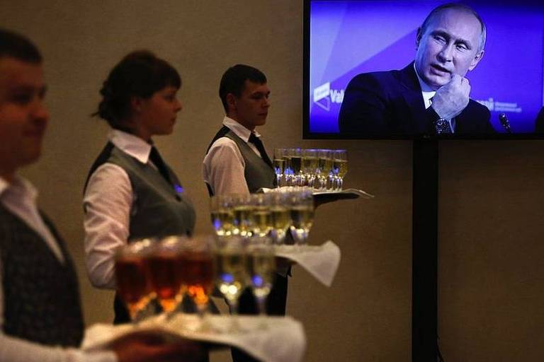 Garçons servem taças enquanto Putin discursa na televisão ao fundo