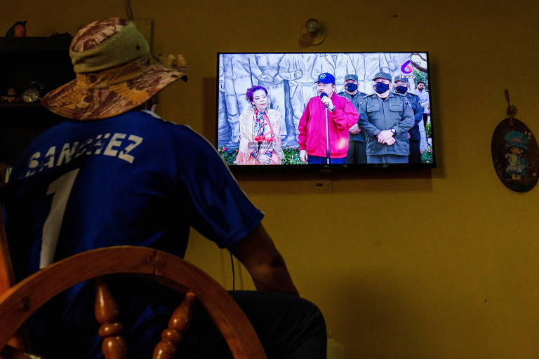 Homem sentado usando chapéu de palha assiste em uma TV a discurso de Daniel Ortega, presidente da Nicarágua, que está acompanhado da primeira-dama e vice-presidente, Rosario Murillo, e de militares