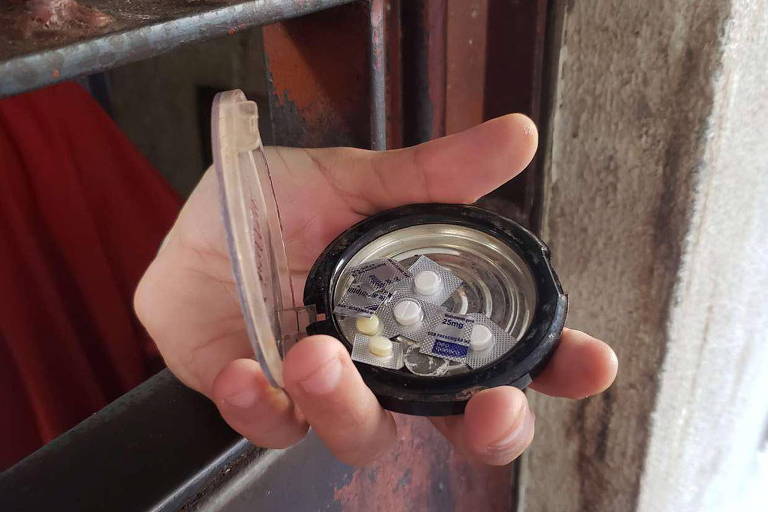 Uma mão segura um recipiente com comprimidos de remédio, a pessoa está dentro de uma cela