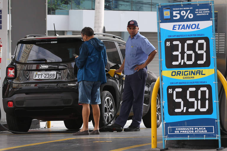 Placa azul com preços de combustíveis em números grandes brancos, emprimeior plano. Ao fundo, frentista colcoa combustível em carro preto