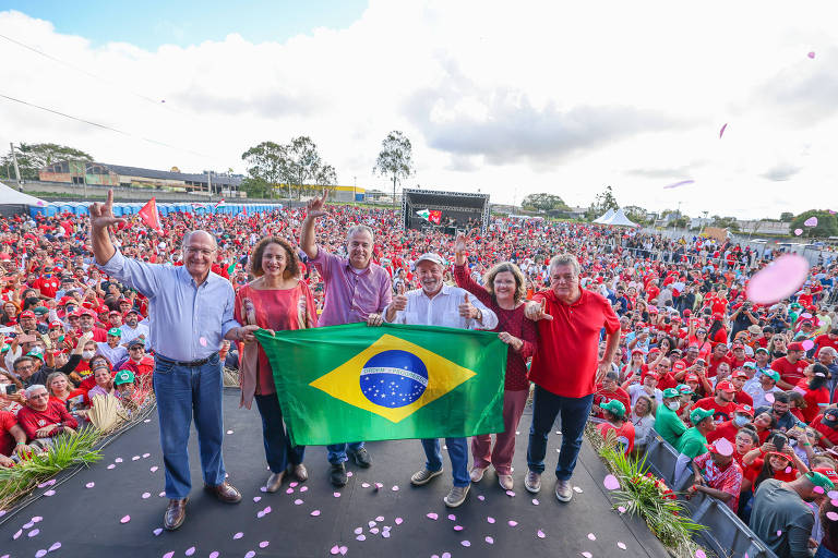 Seis pessoas em pé (Lula entre eles), em palco, segurando bandeira do Brasil, com plateia de vermelho ao fundo