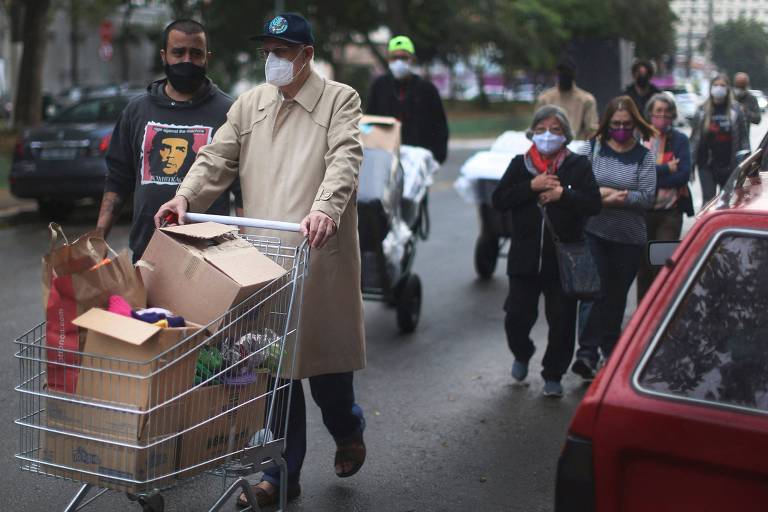 Imagem mostra Padre Júlio empurrando carrinho de compras cheio de caixas. Ele é seguido por algumas pessoas, numa rua.