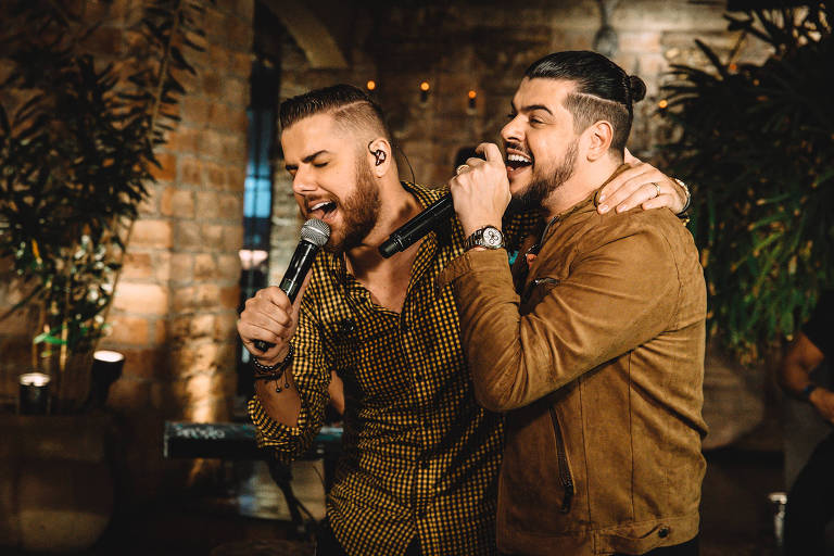Em foto colorida, dois homens se abraçam enquanto cantam em um show