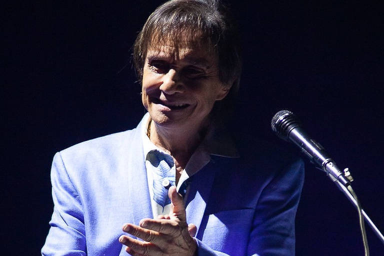 Roberto Carlos de terno azul bate palmas no palco, ao lado do microfone