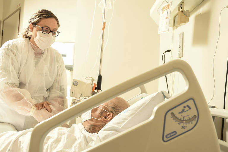 Paciente com leucemia, usando máscara, rdá a mão direita para enfermeira, que a segura com suas duas mãos