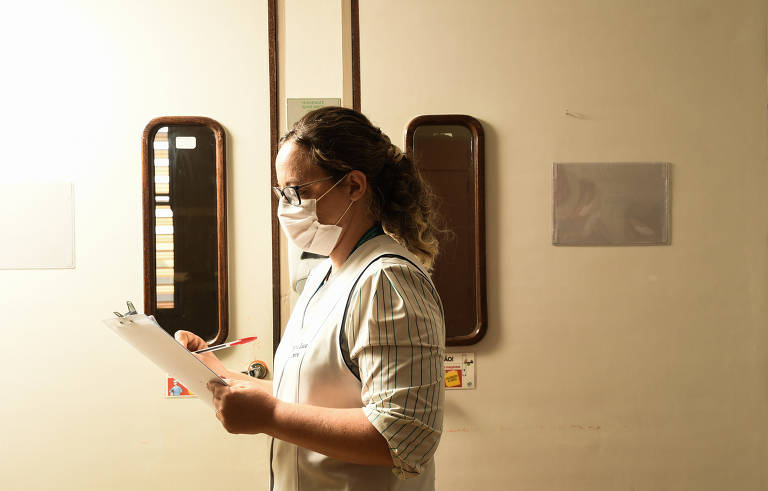 Imagem mostra enfermeira de lado, com máscara e segurando papéis. Ela está na frente de uma porta.