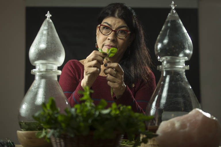 Uma mulher branca com longos cabelos escuros usa óculos e veste uma blusa vinho. À sua frente, em uma mesa, vemos plantas verdes e dois grandes tubos de vidro usados na produção de essências.