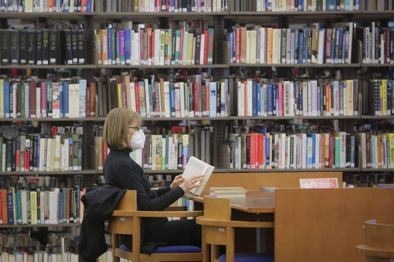 Imagem mostra mulher lendo livro sentada em mesa e perto de estantes cheias de livros