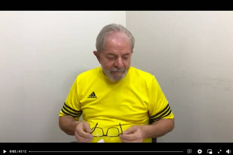 Print de tela de vídeo no Facebook com Lula de camiseta amarela com faixas pretas nas mangas. Ele segura óculos nas mãos. Fundo parede clara