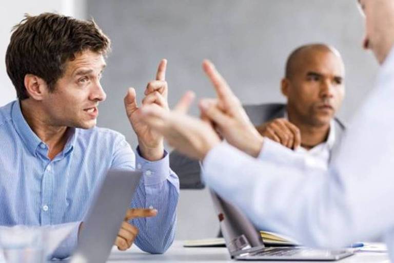 Homens em uma mesa apontam dedos um para o outro sinalizando que estão discutindo