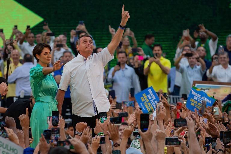 Michelle, de vestido verde, e Bolsonaro levantando a mão esquerda, rodeados por público