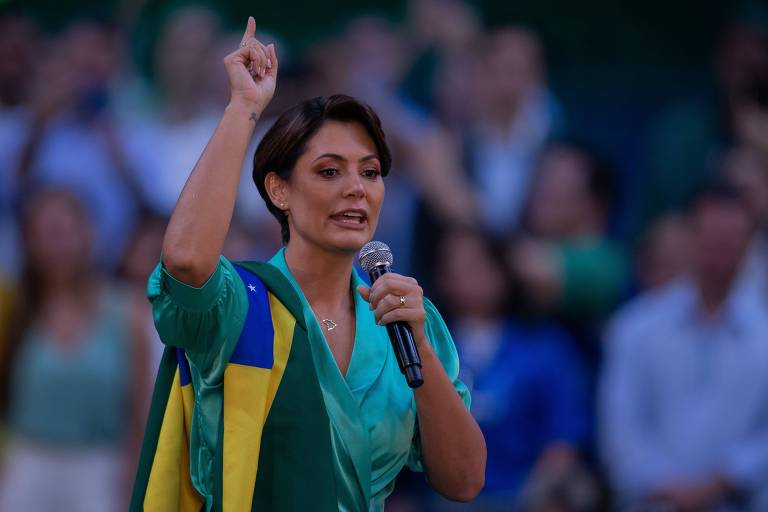 Fotografia colorida mostra primeira-dama Michelle Bolsonaro durante discurso. Ela utiliza um microfone e está com uma bandeira do Brasil pendurada no ombro direito, com o braço levantado. 