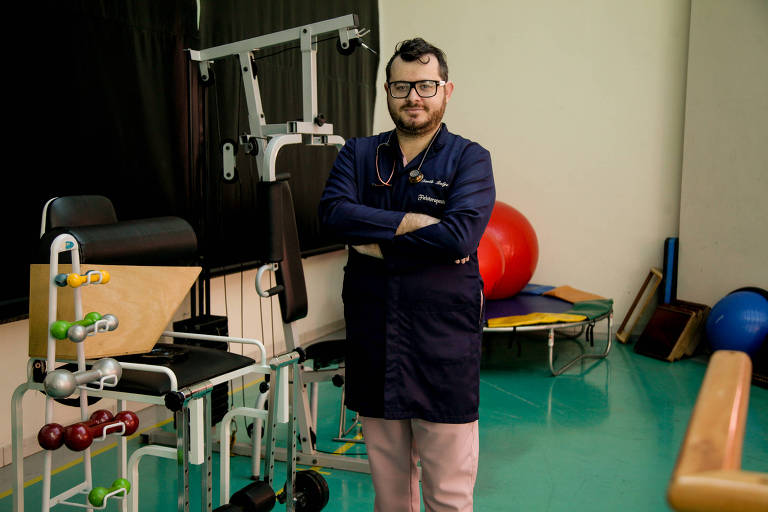 Danilo, um homem branco, de cabelos e barba castanhos, está em uma clínica de fisioterapia; ele está em pé, com os braços cruzados, usando óculos de grau, jaleco azul escuro e calça em um tom claro de cor-de-rosa; ao redor dele, estão equipamentos como halteres e cama elástica