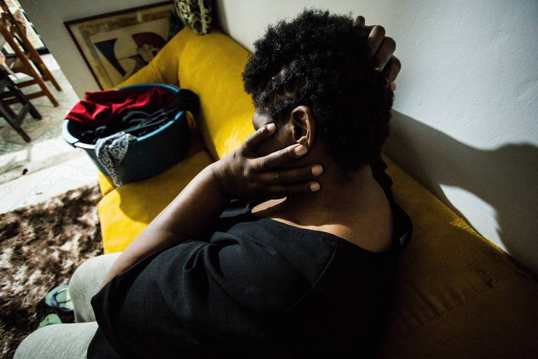 Uma mulher negra está sentada em um sofá, de costas para a câmera, com a mão no rosto. Ela foi vítima de trabalho escravo doméstico