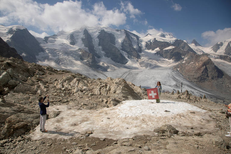 Mulher segura bandeira enquanto homem a fotografa em uma paisagem montanhosa; há neve só no pico das montanhas
