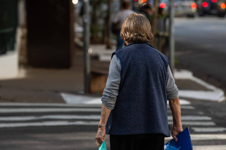 Idosa atravessa a rua na região central de SP; ela usa uma blusa azul com mangas cinza, carrega uma sacola em cada mão, está de costas e tem o cabelo curto e castanho