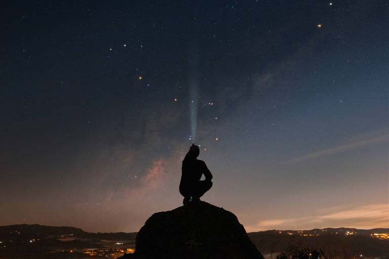 Céu com estrelas, é possível ver a via láctea e a silhueta de um fotógrafo mirando o céu. Ele está sobre uma pedra no Pico do Olho d´água em Mairiporã