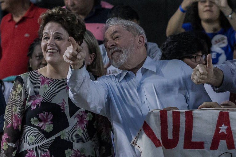 Dilma Rousseff e Lula, que aponta algo com a mão direita para a companheira de PT, estão em um palanque em Ipatinga, em evento na cidade mineira