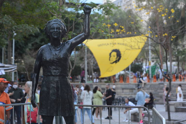Estátua feita em homenagem a Marielle é instalada em praça com um bandeira com o rosto da vereadora assassinada ao fundo