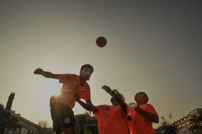 Três homens vestindo camisetas laranja disputam jogada durante treino de futsal. A bola está no ar, após um deles a ter cabeceado 