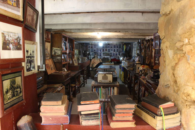 em primeiro plano, estão alguns livros empilhados. um vão na parede mostra uma ampla sala onde estão diversos objetos históricos organizados em estantes e mesas