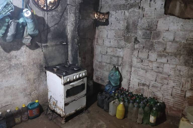 A imagem mostra uma cozinha suja com um fogão com a lateral enferrujada, garrafas de plástico no chão e paredes com reboco de cimento e tijolos sem tinta